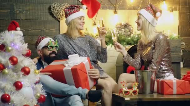 Brodaty mężczyzna siedzi na krześle i posiada wielki prezent, i dwa białe dziewczyny w nowy rok kapelusze pić szampana na tle nowy rok. Koncepcja nowego roku nastroju. — Wideo stockowe