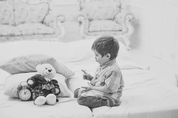 Boy with happy face puts favourite toy on bed, time to sleep. Kid put plush bear near pillows and alarm clock, luxury interior background. Child in bedroom with silence gesture. Time to sleep concept. — Stock Photo, Image