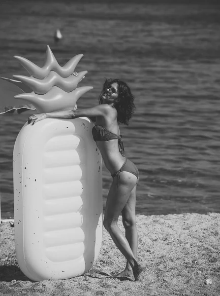 Дама с воздушным матрешкой в форме соснового яблока стоит на пляже в стильном бикини и солнцезащитных очках. Летние каникулы. Женщина держит воздушный матрас ананас в форме моря или океана на заднем плане . — стоковое фото