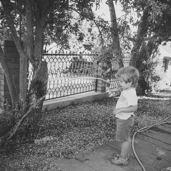 Αγόρι με μάνικα χύνει νερό στο δέντρο ή και θάμνων. Άρδευση φυτών της έννοιας. Παιδιού, παιδί απασχολημένο πρόσωπο βοηθώντας στο πότισμα, ενυδατικό έδαφος. Μικρός βοηθός στο κήπο κατέχει σωλήνα με ροή νερού. — Φωτογραφία Αρχείου