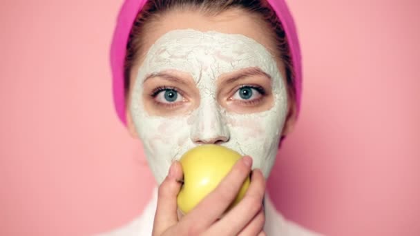 Nahaufnahme eines Mädchens mit Maske im Gesicht, das einen Apfel auf rosa Hintergrund isst. Konzept der Hautpflege und Gesichtsreinigung. Konzept eines gesunden Lebensstils. — Stockvideo