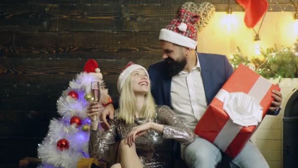 Hombre barbudo con una gran caja de regalo se sienta cerca de una hermosa chica con una copa de champán en la mano en el fondo del año nuevo. Una pareja cariñosa con un regalo de Año Nuevo. Año Nuevo amor . — Vídeos de Stock