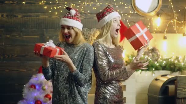 Mädchen freuen sich über Neujahrsgeschenke. erstaunt überraschte glückliche blonde Mädchen in silbernen Kleidern und roten Hüten mit Neujahrsgeschenken in der Hand im weihnachtlichen Atelierraum mit Neujahrsbaum. — Stockvideo