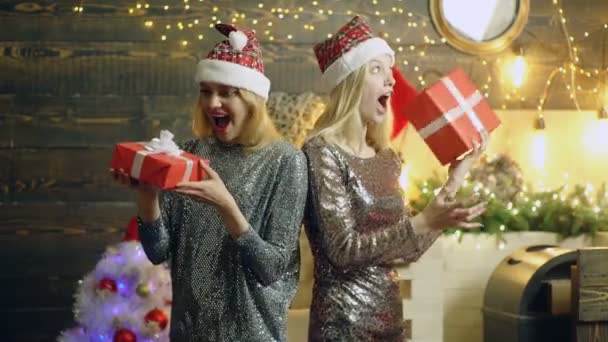 Girls rejoice at New Years gifts. Amazed surprised happy blonde girls in silver dresses and red hats holding New Years gifts in their hands in christmas studio room with new year tree. — Stock Video