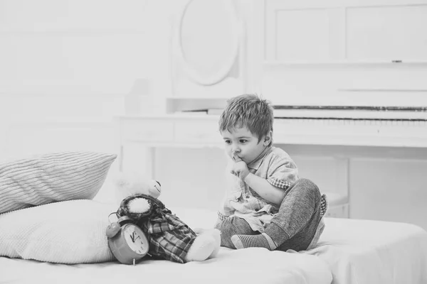 Baby playing bear playing on white bed. Kid put toy in bed for sleep, shows gesture to be silent. Teddy bear sleeps near alarm clock.