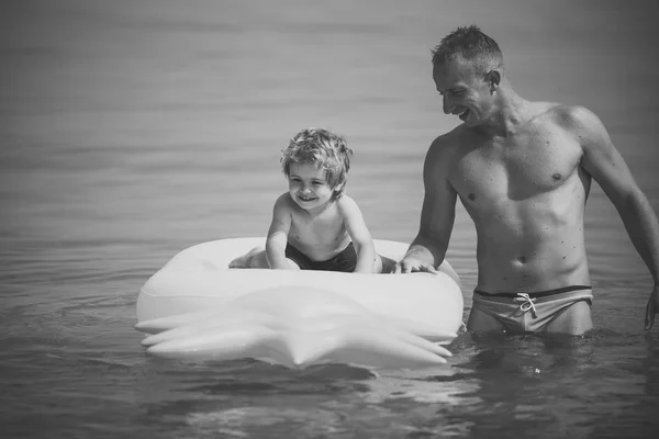 父亲的概念。父亲站在附近, 与儿子推床垫。爸爸和带着笑脸的孩子在海里共度时光。可爱的儿童男孩与坐在空气床垫菠萝形状, 在海洋, 海. — 图库照片