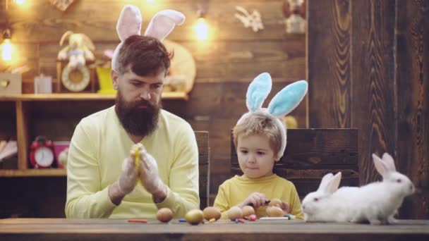 El hombre barbudo y su hijo vestidos con orejas de conejo adornan los huevos de Pascua. Padre e hijo están pintando huevos. La familia feliz se está preparando para la Pascua. Lindo niño con orejas de conejito. Feliz Pascua. . — Vídeos de Stock