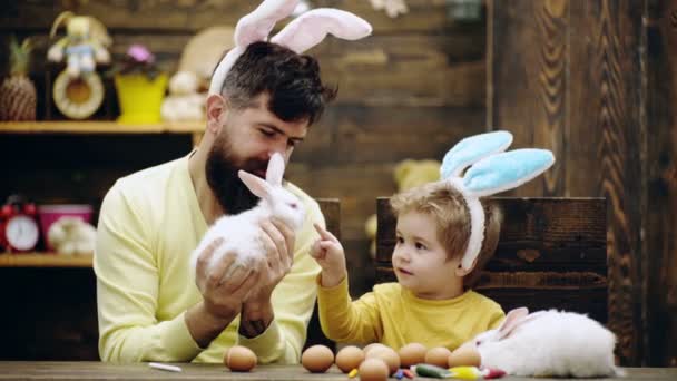 Garçon avec des oreilles de lapin regardant petit lapin moelleux. L'homme tient le lapin de Pâques. Une famille heureuse se prépare pour Pâques. Œufs de Pâques sur fond en bois. Lapin de Pâques . — Video
