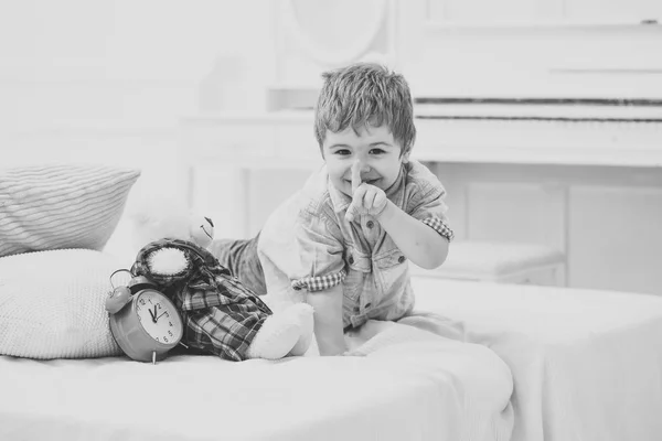 Time to sleep concept. Child in bedroom with silence gesture. Boy with happy face puts favourite toy on bed, time to sleep. Kid put plush bear near pillows and alarm clock, luxury interior background.