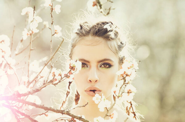 Pretty girl or woman with fashionable makeup on cute face, has stylish hair, posing among blooming spring flowers or cherry blossom white color on tree branch sunny day outdoor on natural background