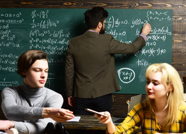 Lehrer mit Brille lesen Berichte talentierter Schüler, die Fehler korrigieren. Aufmerksame Schüler schreiben etwas in ihre Notizblöcke, während sie im Klassenzimmer am Schreibtisch sitzen. — Stockfoto