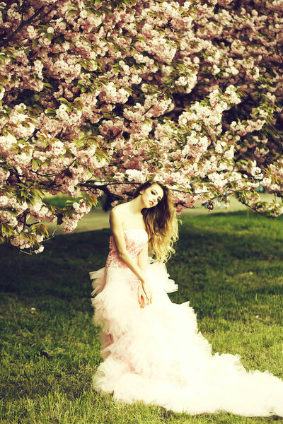 Beautiful young woman in pink glamour dress with long curly hair in spring flowers bloom