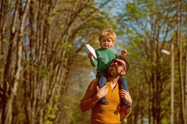 Padre dar hijo a cuestas lanzamiento avión de papel de juguete. Niño pequeño juega con juguete en el parque. Arriba, arriba y lejos — Foto de Stock