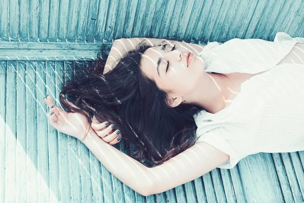 Warmte concept. Vrouw brunette koelen terwijl staat in de buurt van jalousie, strepen van zonlicht en schaduw op haar gezicht. Lady draagt unbuttoned shirt. Meisje op kalme gezicht leunen op turquoise jalousie. — Stockfoto