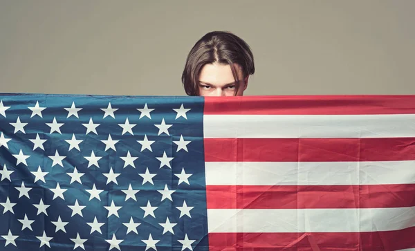 Un hombre con cara seria sostiene la bandera de EE.UU., escondiéndose detrás de la bandera. Un tipo escondido en EE. UU., América lo protege, fondo gris. Concepto de privacidad y conspiración . — Foto de Stock