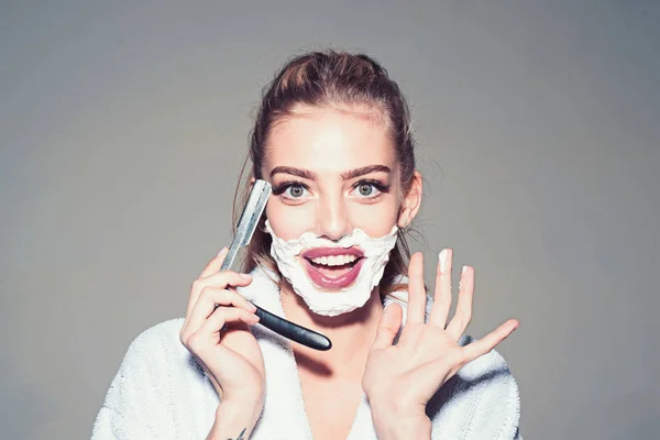 Girl on smiling face wears bathrobe, grey background. Lady play with sharp blade of straight razor. Woman with face covered with foam holds straight razor in hand. Barber and shaving concept. — Stock Photo, Image