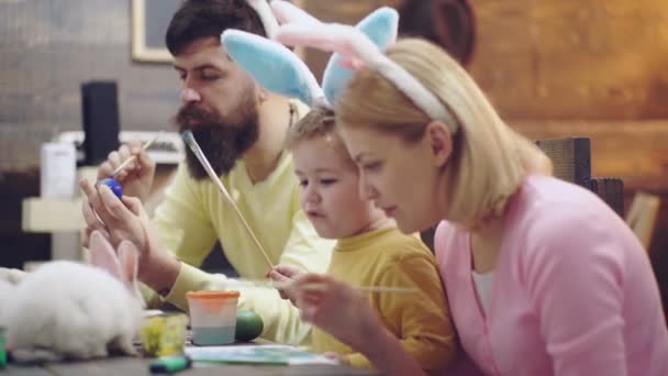 El chico está pintando huevos. Madre, padre e hijo están pintando huevos. La familia feliz se está preparando para la Pascua. Lindo niño pequeño con orejas de conejo . — Vídeos de Stock