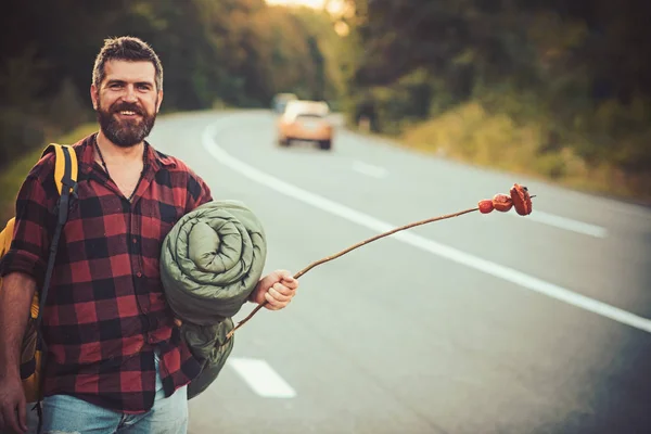 Homem sorridente barbudo com mochila, dormindo de volta segurando pau com salsichas nele. Bonito carona no caminho de volta para casa, wanderlust e conceito de aventura — Fotografia de Stock