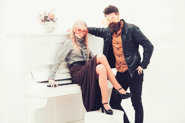Blond girl sitting on white piano while bearded man in leather jacket standing next to her. Couple in enormous orange glasses posing in white studio — Stock Photo, Image