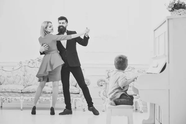 Musician education concept. Boy adorable try to play piano musical instrument, while parents dancing. Child sit next to piano, play music, white interior background. Rich parents enjoy parenthood. — Stock Photo, Image