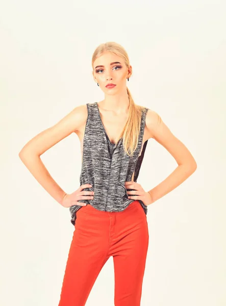 Skinny model with thick eyebrows and neat ponytail wearing melange gray sleeveless blouse and bright orange pants isolated on white background