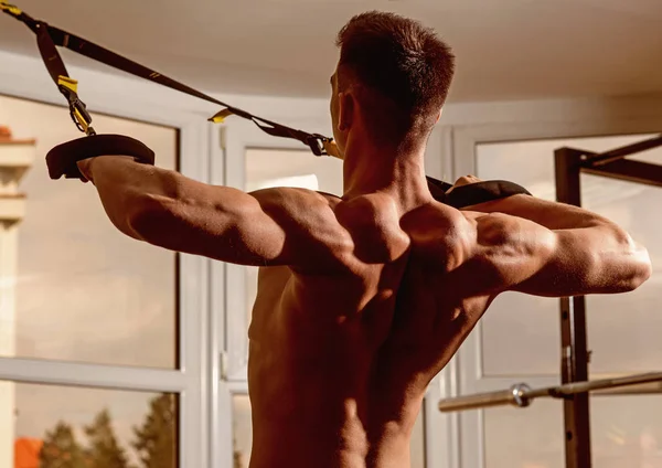 Deportista, atleta, macho muscular hace ejercicio con bucles trx, ventana en el fondo, de cerca. Hombre con torso desnudo, y musculoso de vuelta en el gimnasio disfrutar del entrenamiento, trx. Concepto de deporte y gimnasio . — Foto de Stock