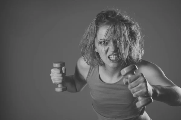 Woman coach with angry face holds dumbbells n grey background.