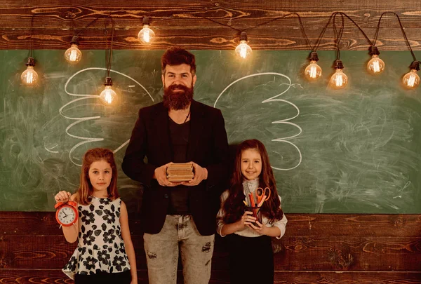 Teacher and girls pupils in classroom, chalkboard on background. Children and teacher with drawn by chalk wings. Man with beard and schoolgirls with school attributes. Favourite teacher concept.