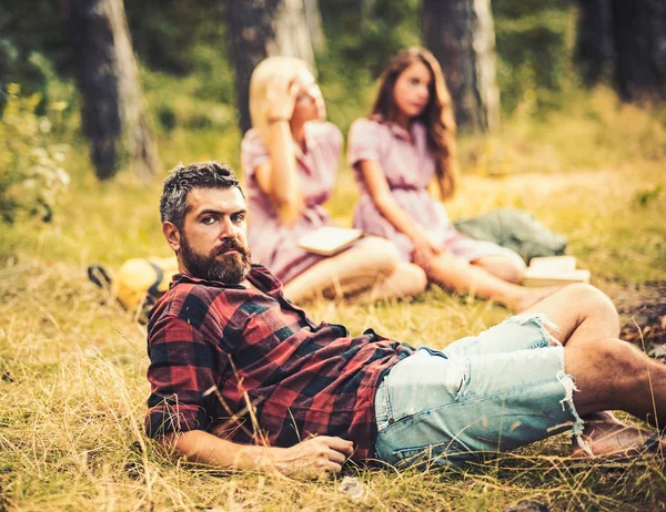 Bearded man in lumberjack shirt spending time with friends outdoors. Handsome man lying on glass in woods. Outdoor recreation and nature concepts