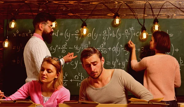 Students, group mates studying, while teacher asking girl near chalkboard. Exam concept. Bearded teacher, , professor asking student to solve mathematical equation chalkboard on background