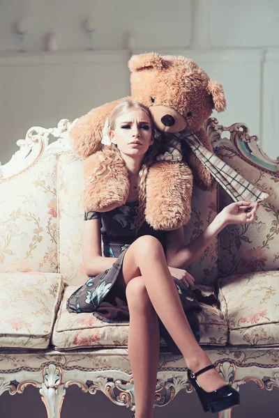 Молодой избалованный аристократ сидит на винтажном диване. Блондинка играет со своим огромным плюшевым мишкой — стоковое фото