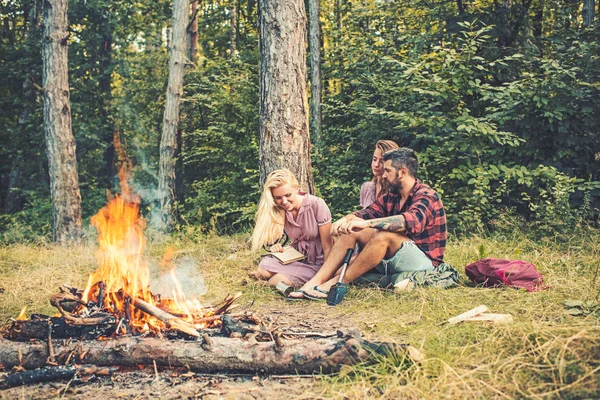 Active lifestyle concept. Group of friends camping in the forest. Girl reading next to bonfire