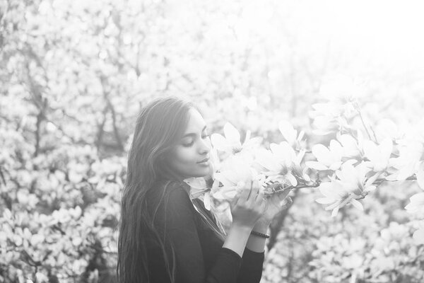 Adorable girl or pretty woman with brunette, long hair enjoying pink, magnolia flower blossom on trees in flowering park on sunny day on blurred floral environment. Daydreaming. Flourishing and spring