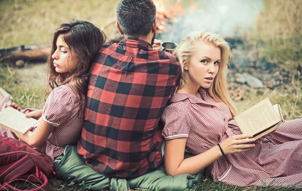Boyfriend and girlfriends read books. Man with women at bonfire. Friends enjoy camping vacation. People relax at fire on summer nature. Love triangle, cheating and relations