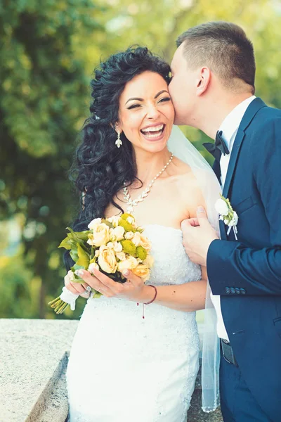 Groom embrasser mariée heureuse avec bouquet. Femme et homme sourient le jour du mariage. Couple amoureux de mariage. Couple nouvellement marié en été en plein air. Concept de mariage. amour et famille — Photo