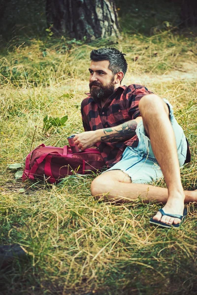 Турист в клетчатой рубашке держит кружку. Хипстер с длинной бородой отдыхает на зеленой траве. Бородатый мужчина с чаем или чашкой кофе в лесу. Кемпинг, выпивка и кофе настроение. Летние каникулы — стоковое фото