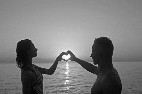 Woman and man with heart hands at sea water in sunset. Summer holidays and travel vacation. Family and valentines day. Couple in love relax on sunrise beach. Love relations of couple enjoying summer. — Stock Photo, Image
