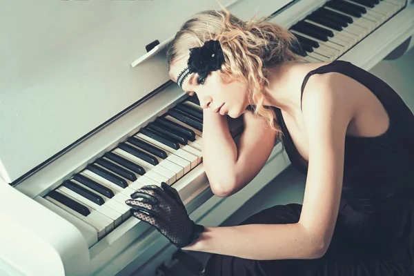 Junge Schauspielerin am Klavier liegend, überwältigt von Erinnerungen. trauriges blondes Mädchen, das sich an die Tastatur lehnt. Modekonzept der wilden Zwanziger — Stockfoto