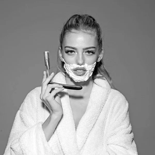 Chica en la cara tranquila lleva albornoz, fondo gris. Mujer con la cara cubierta de espuma sostiene la navaja recta en la mano. Señora jugar con la cuchilla afilada de afeitar recta. concepto de peluquero y afeitado . — Foto de Stock