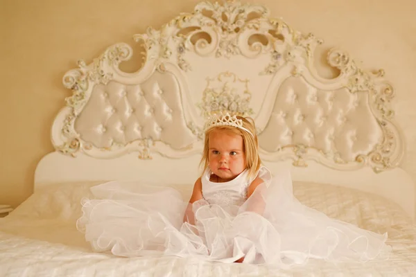 Справжня принцеса. Аксесуар для волосся. Маленька дівчинка носить корону і зачіску. Маленька дитина з довгим світлим волоссям. Маленька блондинка сидить на ліжку. Красиві зачіски для дівчаток. Її краса сяє через — стокове фото