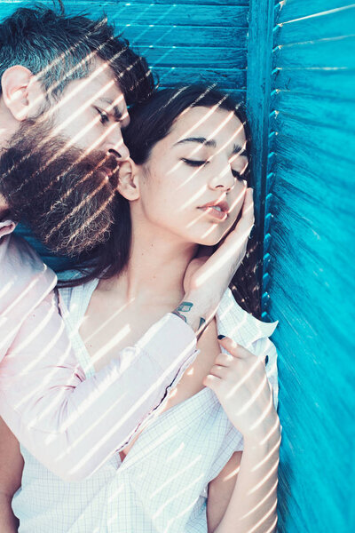 Heat concept. Couple chilling near jalousie, stripes of sunlight and shadow on faces. Girl in unbuttoned shirt and man with beard. Couple in love on calm faces near turquoise jalousie.