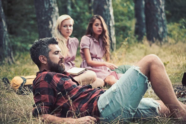 Man camping in forest with women on blurred background. Man with beard relax on grass.