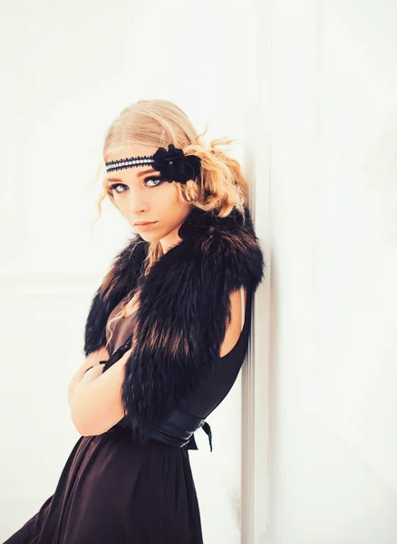 Young movie star of roaring twenties posing isolated on white background. Blond girl in headband with flower and fur collar leaning on wall — Stock Photo, Image