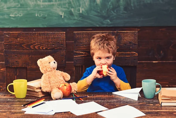 School break. Hungry kid biting apple in classroom. Small boy playing with paper plane and teddy bear