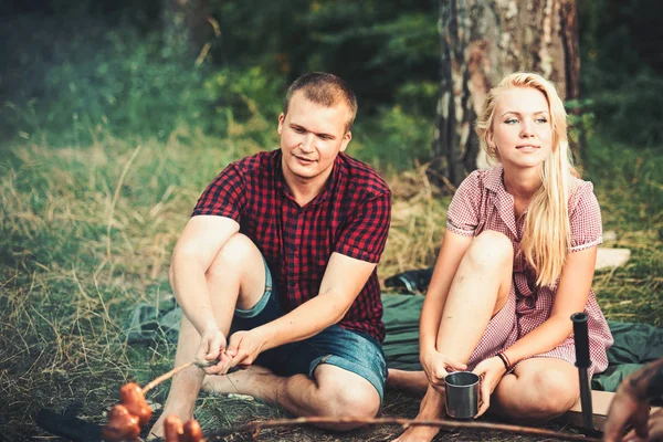 Schönes verliebtes Paar am Lagerfeuer sitzend. romantisches Date in der Natur. blondes Mädchen in schönem Retro-Kleid trinkt Tee, während ihr Freund Essen kocht — Stockfoto