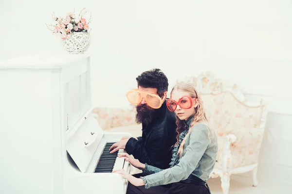 Pareja joven en enormes vasos sentados en habitación blanca y tocando el piano. Elegante hombre de cuentas y su novia rubia sentado en el piano de cola — Foto de Stock