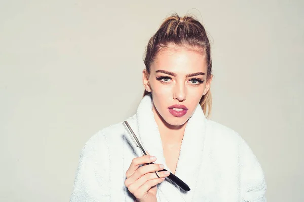 Girl on calm face wears bathrobe, grey background. Woman with attractive lips holds straight razor in hand. Barber concept. Lady play with sharp blade of straight razor