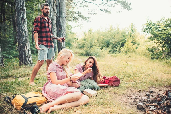 Women read book at campfire. Friends relax at fire. People at bonfire on green grass. Summer vacation concept. Camping, hiking and travel