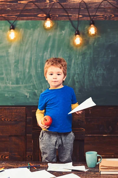 Mały chłopiec trzyma papierowy samolot i apple w jego ręce. Uczeń w niebieski T-shirt stojącą za tabeli. Koncepcja edukacyjna gra — Zdjęcie stockowe