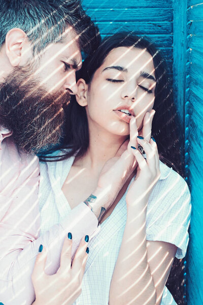 Couple chilling near jalousie, stripes of sunlight and shadow on faces. Heat concept. Couple in love on calm faces near turquoise jalousie. Girl in unbuttoned shirt and man with beard.